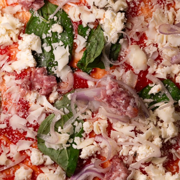 Uncooked pizza with tomato sauce, basil, red onion, crumbled feta, and ground sausage.