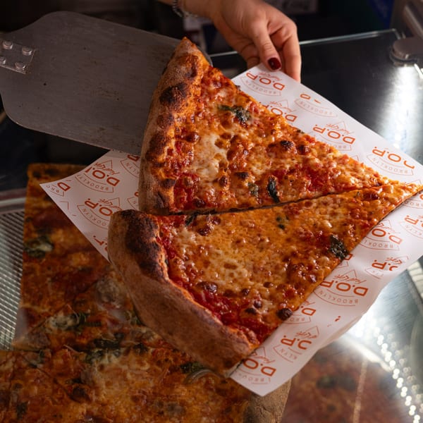 Two slices of Brooklyn DOP FastLife cheese pizza on branded paper, held by a hand.