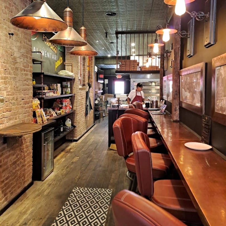 Interior of Brooklyn DOP FastLife, with a long bar, brown leather stools, and a chef in the kitchen.