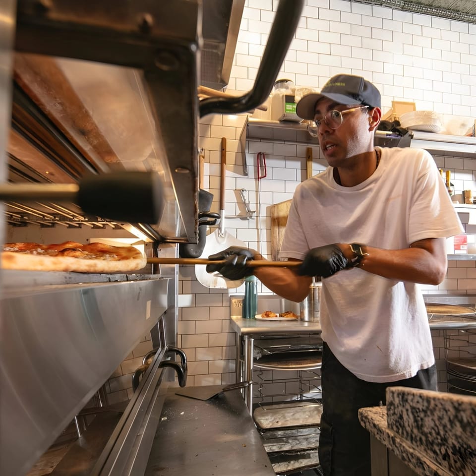 A Brooklyn DOP FastLife chef in a cap and glasses removes a pizza from a large oven.
