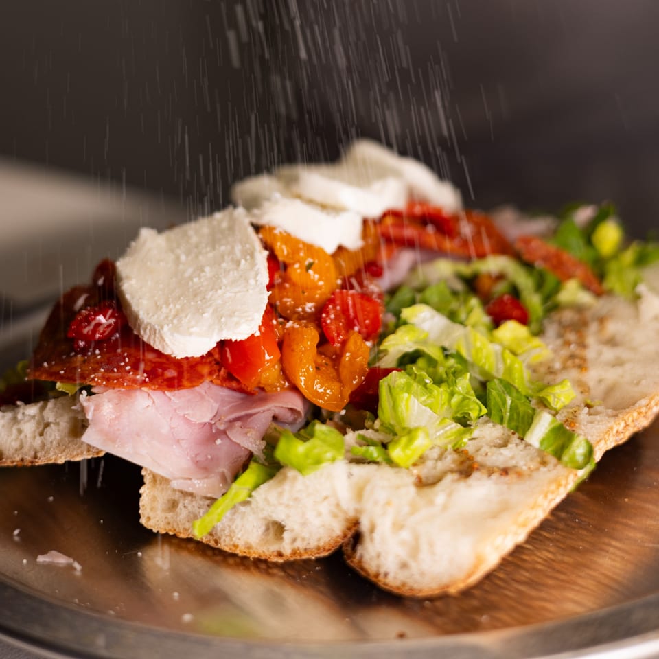 A Brooklyn DOP FastLife sandwich with ham, salami, mozzarella, lettuce, and peppers, being sprinkled with cheese.