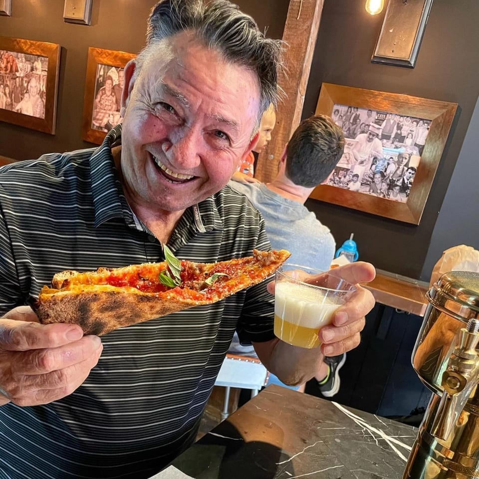 A smiling man holds a slice of Brooklyn DOP FastLife pizza and a drink.
