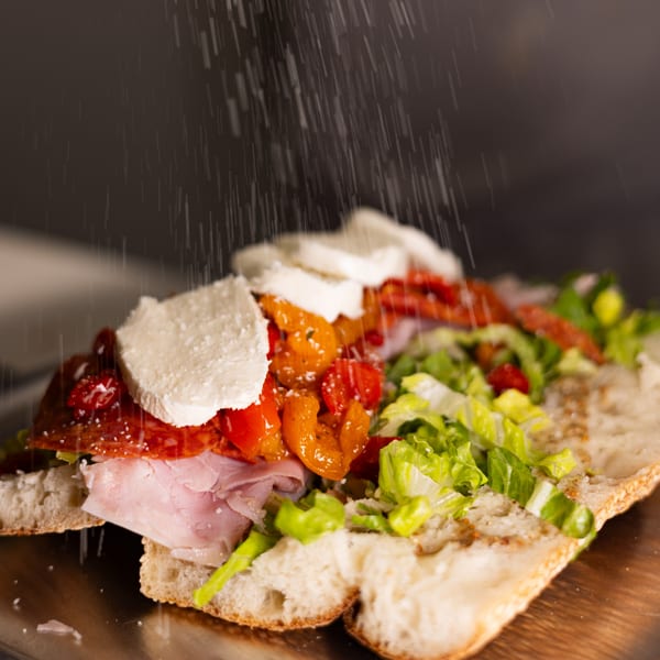 Close-up of a Brooklyn DOP FastLife sandwich with cheese, meats, and vegetables, being sprinkled with cheese.