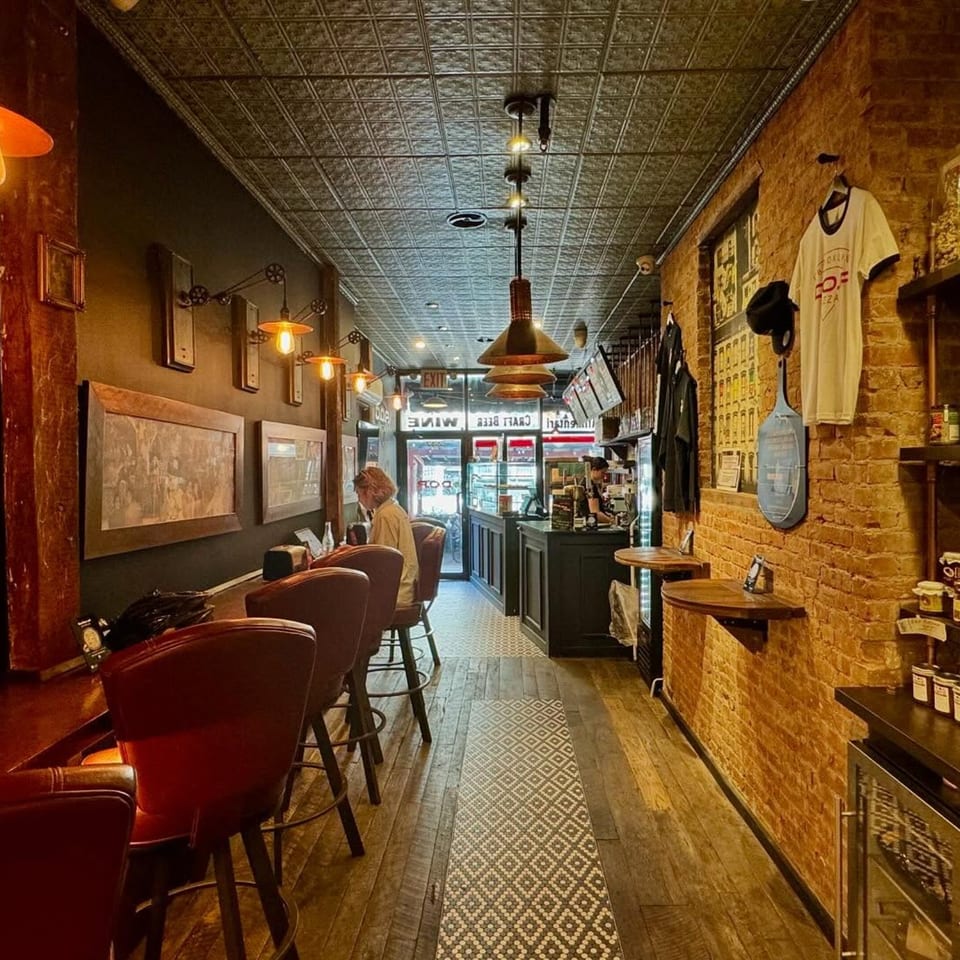 Interior of Brooklyn DOP FastLife, a narrow pizza restaurant with brick walls, bar seating, and a person at the counter.
