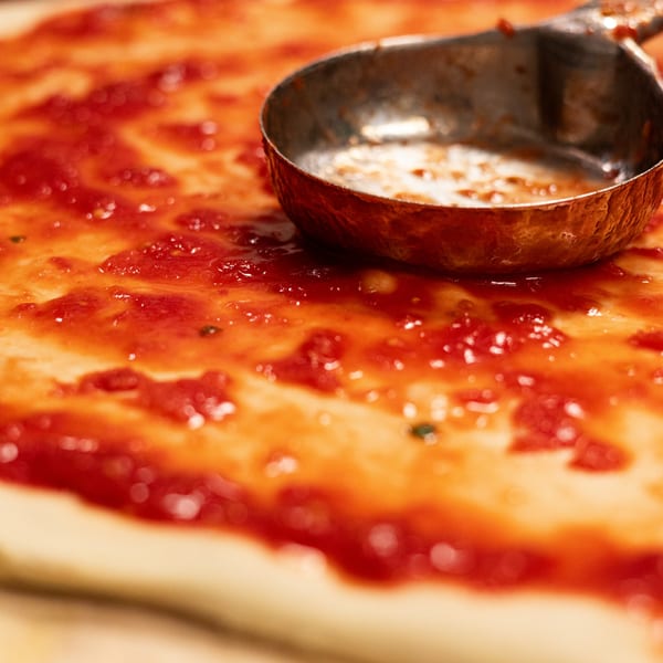 A metal ladle spreading bright red tomato sauce on a pizza dough at Brooklyn DOP FastLife.