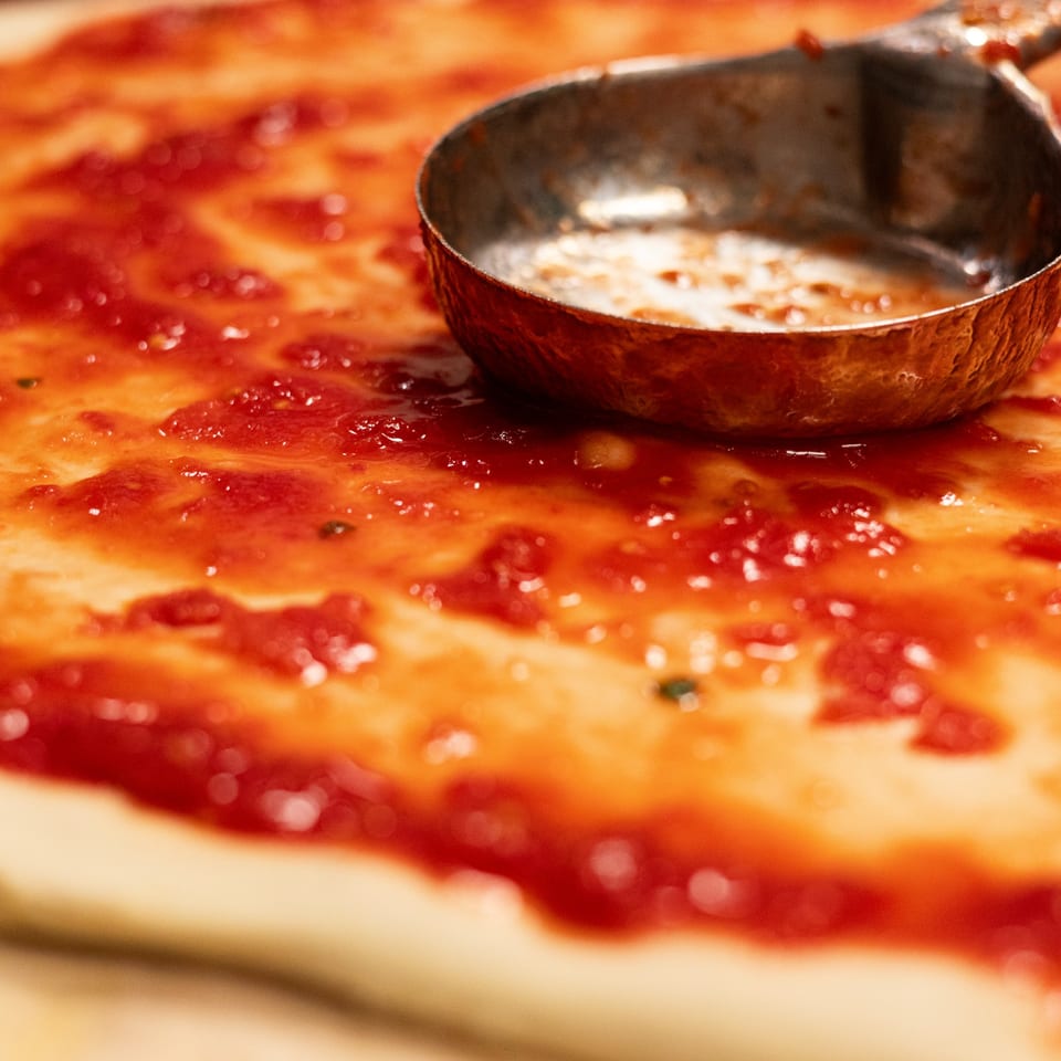 A metal ladle spreading bright red tomato sauce on a pizza dough at Brooklyn DOP FastLife.